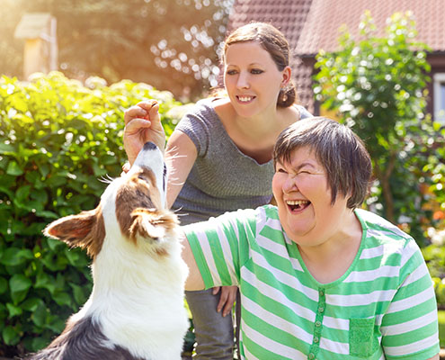 Mentally disabled woman outdoors in the summer, interacting with a companion dog while her female caretaker guides the interaction.