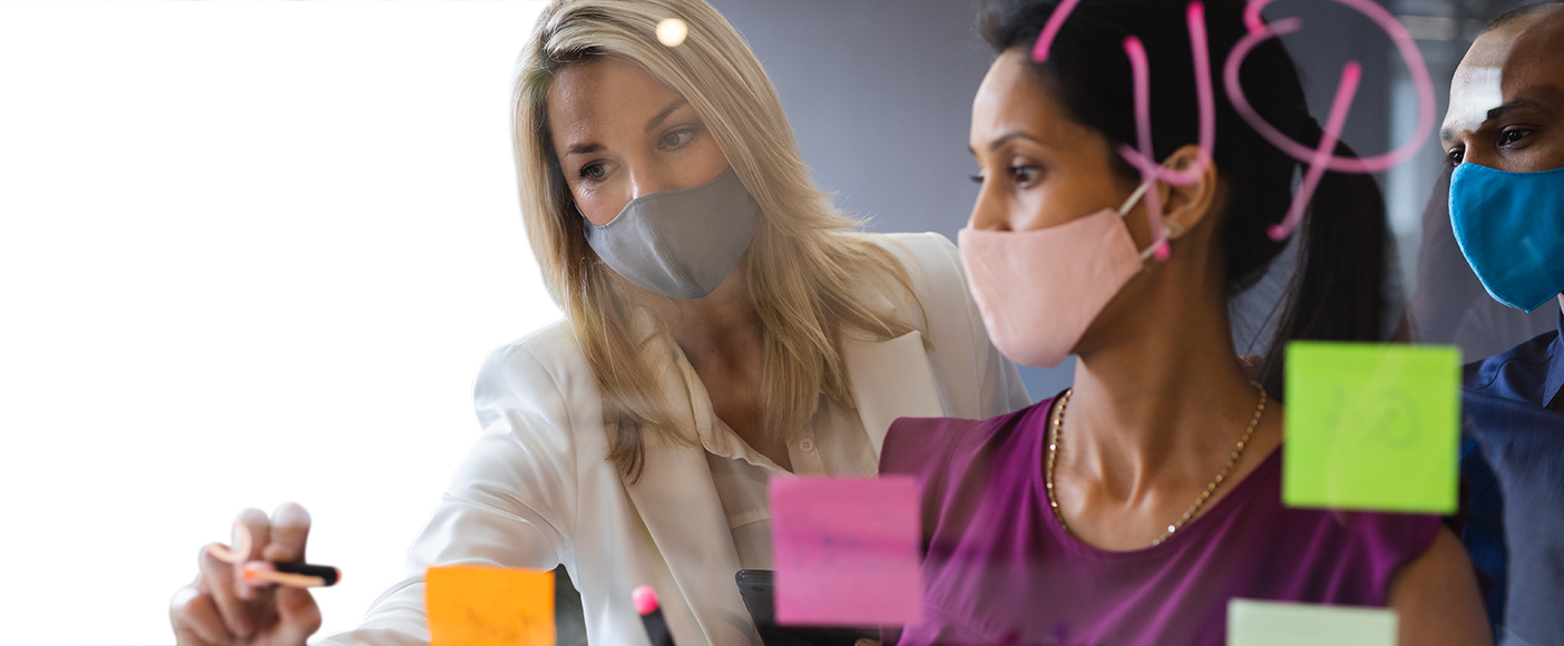 Diverse businesspeople wearing face masks and brainstorming by writing and using sticky notes on a glass board in the office.
