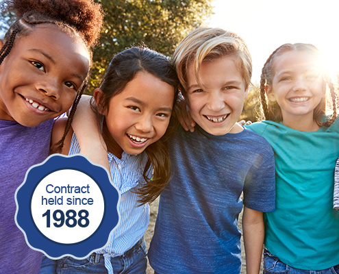 Four diverse, happy, school-age children huddle together for a close-up portrait. A graphic reading, "Contract held since 1988" is added to the image.