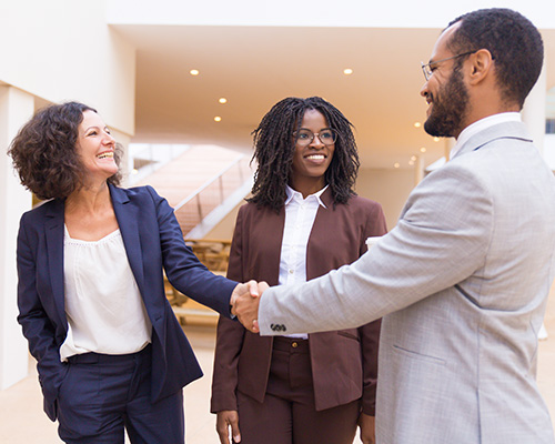 Diverse happy business partners wearing formal business attire greet each other by shaking hands in a lobby area.