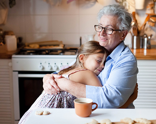 A senior woman sitting in her kitchen warmly embracing a school-age child.