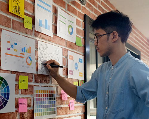 Young Asian male creative thinking while writing a plan on a piece of paper, which is taped up to a brick wall along with other brainstorming papers and sticky notes.