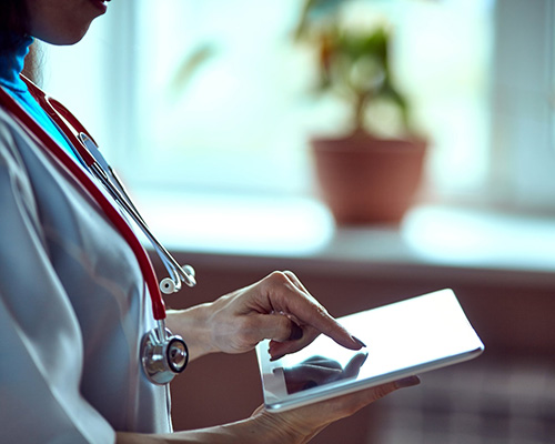 A close-up of a female physician wearing a stethoscope and inputting data into a tablet that she’s holding.