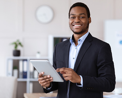 A professionally dressed smiling young Black man holds a tablet in a casual, well-lit office setting.