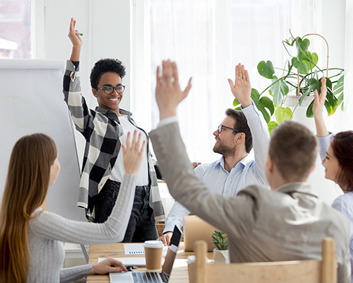 Diverse business people raise their hands while listening to a cheerful consultant standing at the head of the table leading the meeting.