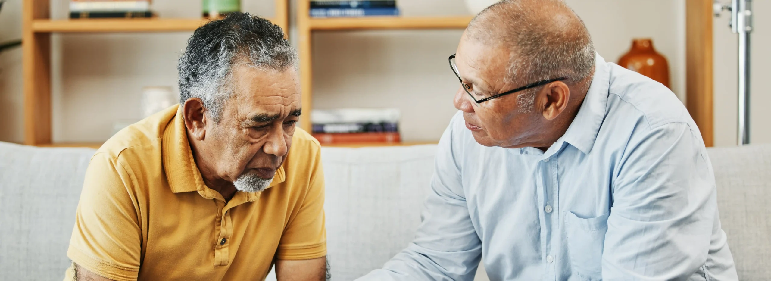Healthcare worker sits on a couch next to an older man, offering support.