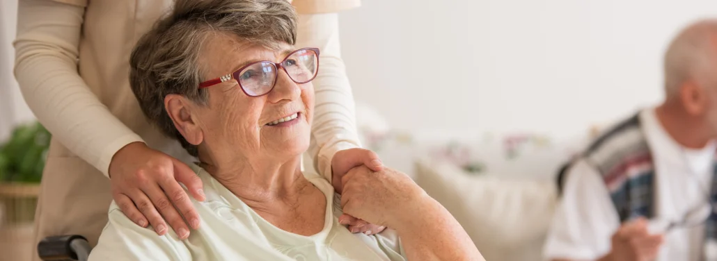 An older woman sits while a woman standing behind her holds her hand.
