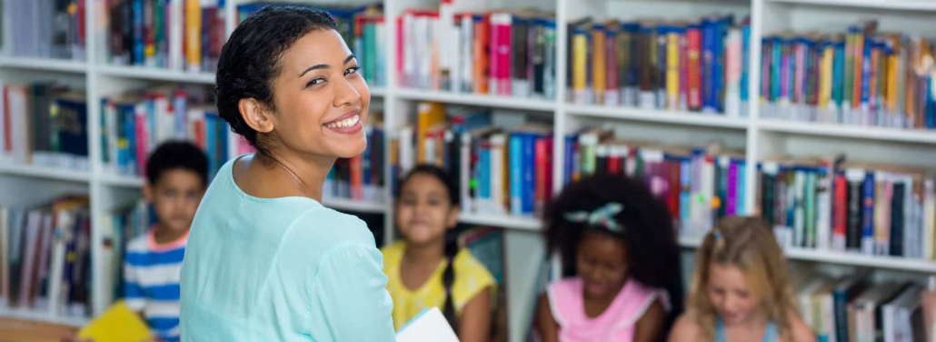 Smiling educator stands in a library holding a book while young children sit together reading in the background.