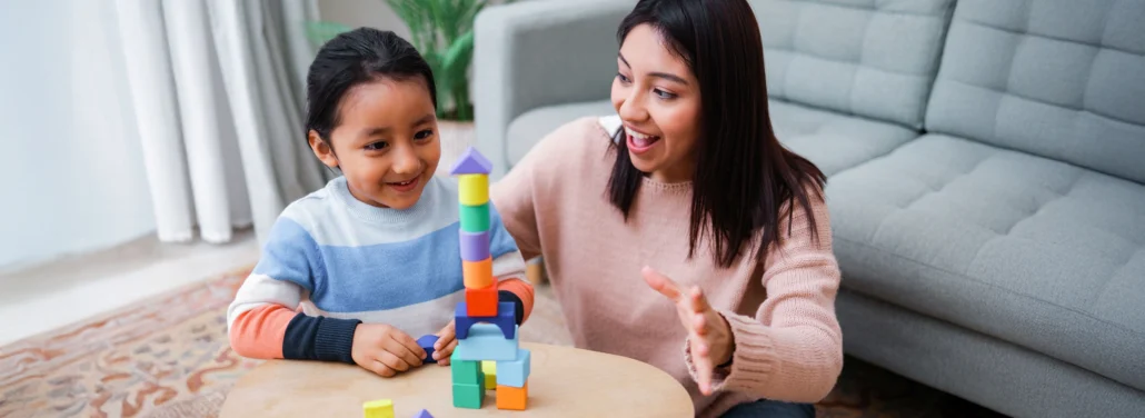 Adult and young child smile while stacking colorful wooden blocks together on a small table.