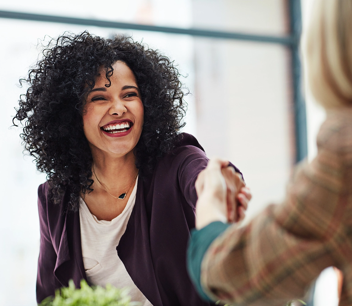 A happy woman in business casual attire shakes hands with another person.